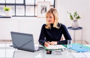 A blonde-haired woman sitting at the desk in the office and working. She is wearing a black turtleneck sweater and using a laptop.